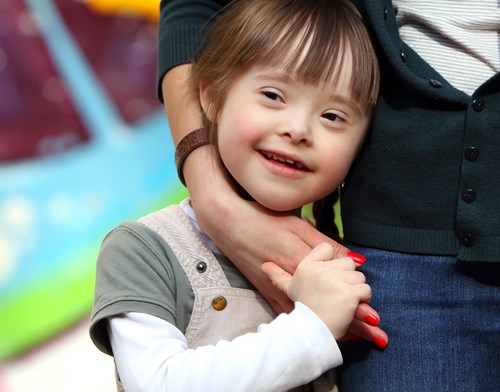 Young child with Downs Syndrome standing next to and holding the hand of her foster carer