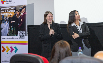 Two women stand in front of a screen, each holding a microphone, engaged in a discussion or presentation.