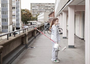 girl standing outdoors on a balcony of run down flats holding a paint roller and tin