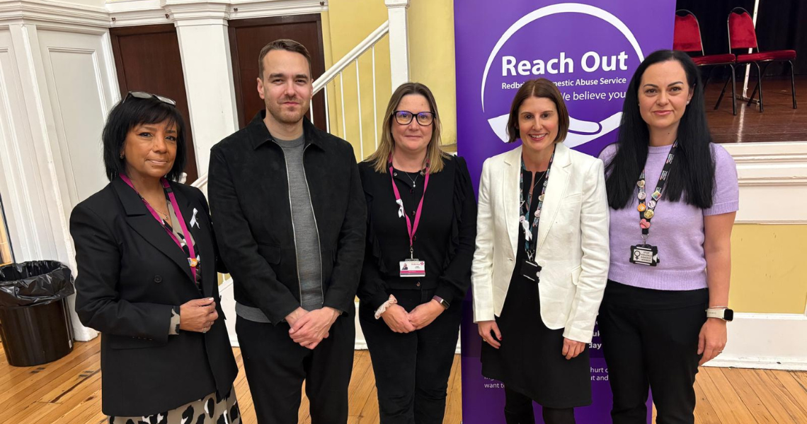 Five people stand in front of a banner for Redbridge Domestic Abuse Service at an indoor event.