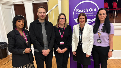 Five people stand in front of a banner for Redbridge Domestic Abuse Service at an indoor event.