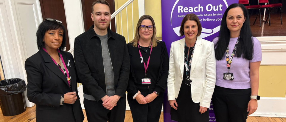 Five people stand in front of a banner for Redbridge Domestic Abuse Service at an indoor event.