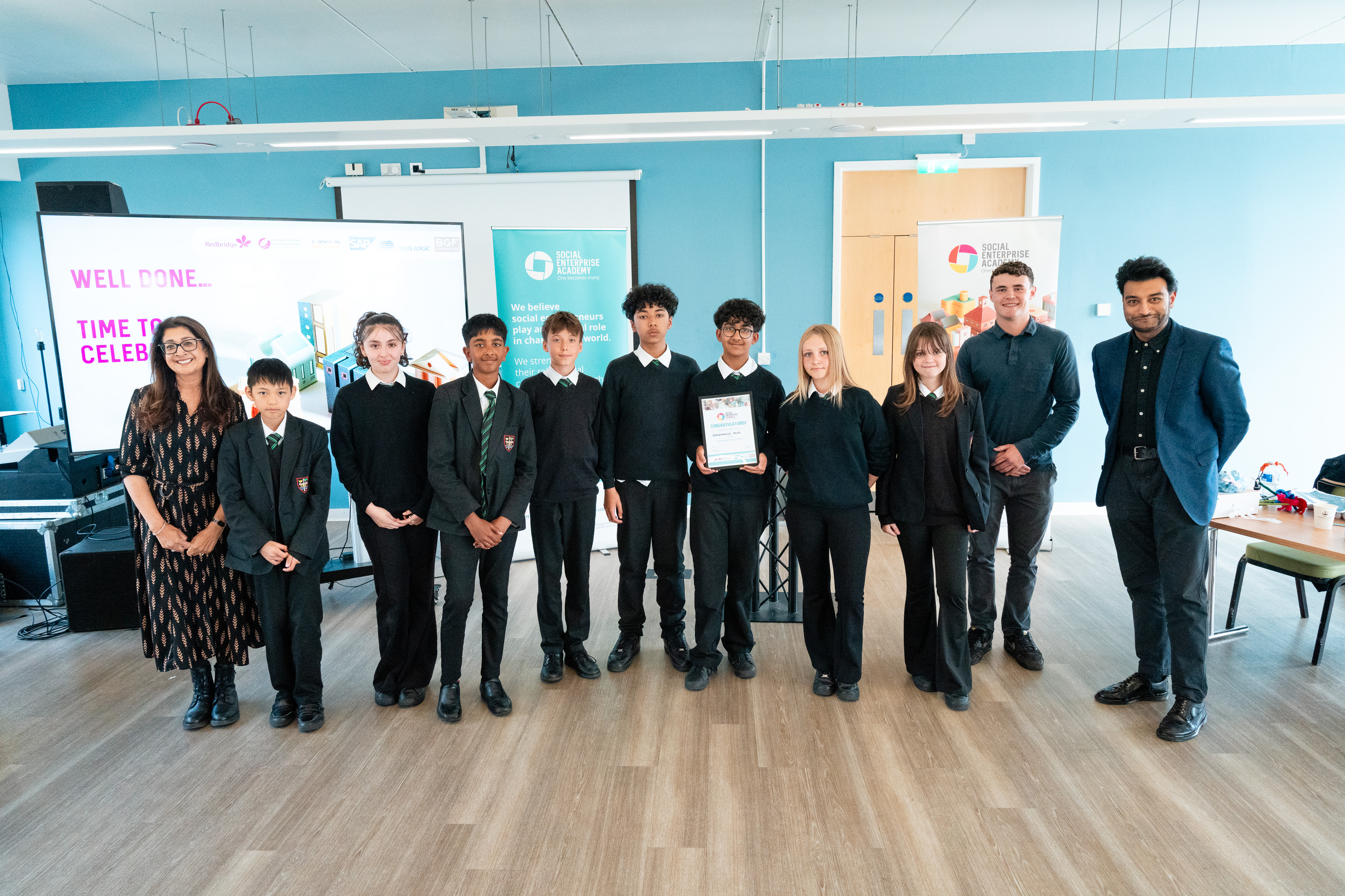   A group of eleven people stand together in a bright room with light blue walls and wooden flooring. The group includes several students in school uniforms and a few adults in professional attire.