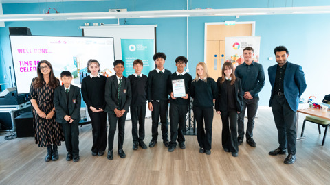   A group of eleven people stand together in a bright room with light blue walls and wooden flooring. The group includes several students in school uniforms and a few adults in professional attire.