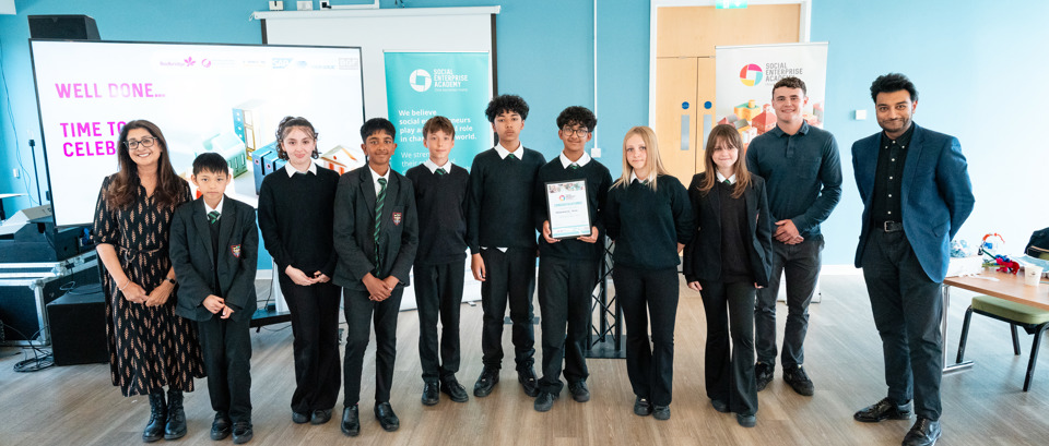   A group of eleven people stand together in a bright room with light blue walls and wooden flooring. The group includes several students in school uniforms and a few adults in professional attire.