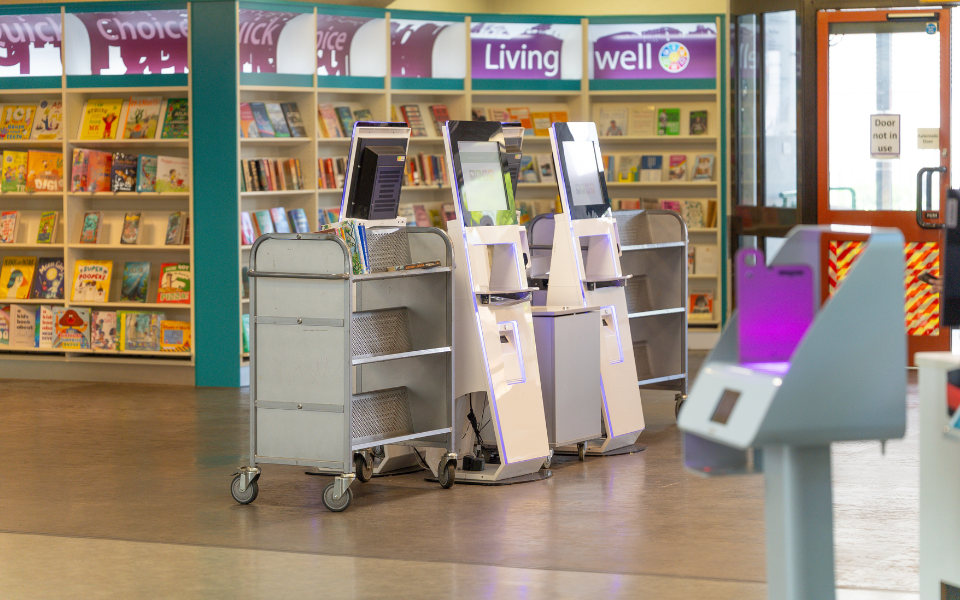 Three self-service checkout kiosks and two book carts are positioned in a library area with bookshelves and magazines visible in the background.