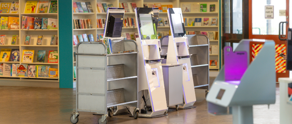 Three self-service checkout kiosks and two book carts are positioned in a library area with bookshelves and magazines visible in the background.