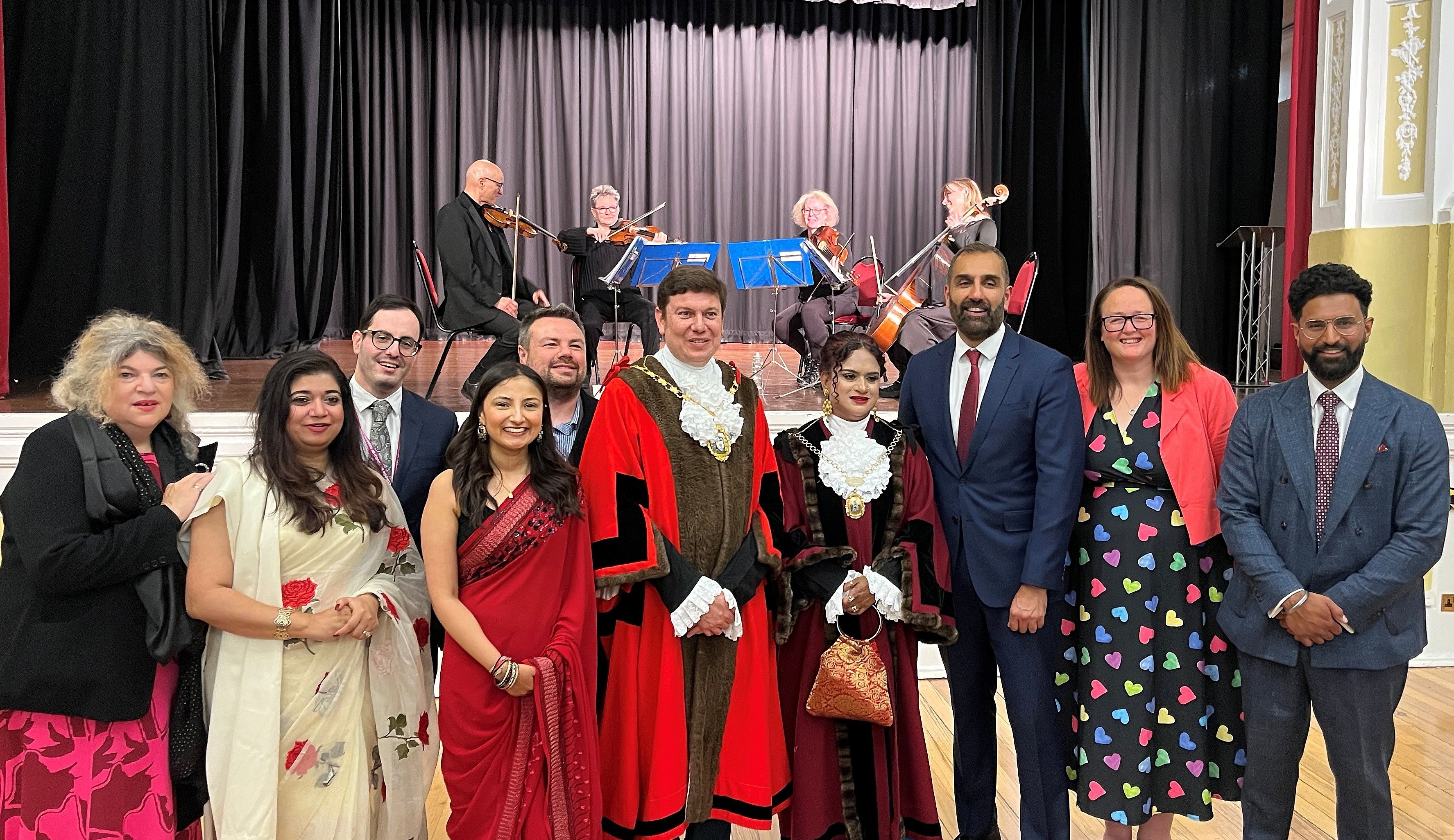 A group of Cllrs in formal attire, including traditional and modern dress, pose in front of a stage with musicians, conveying a celebratory mood.