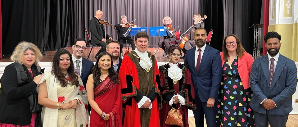 A group of Cllrs in formal attire, including traditional and modern dress, pose in front of a stage with musicians, conveying a celebratory mood.