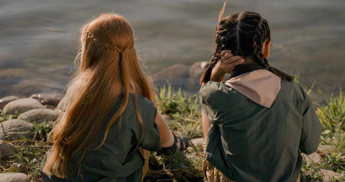 Two girls of primary school age sitting by a lake, taken from the back