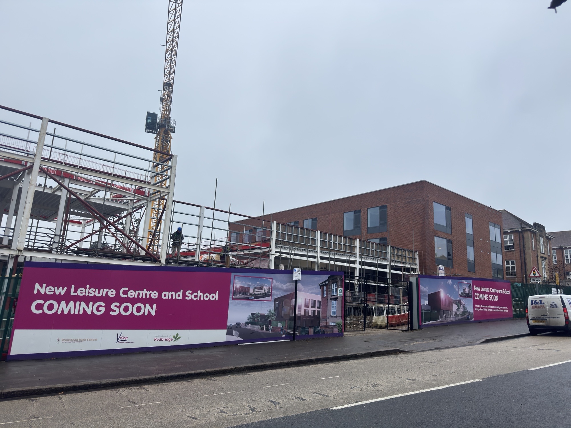 Construction site with a red brick building partly completed, surrounded by a fence with “New Leisure Centre and School Coming Soon” signage. Overcast sky.