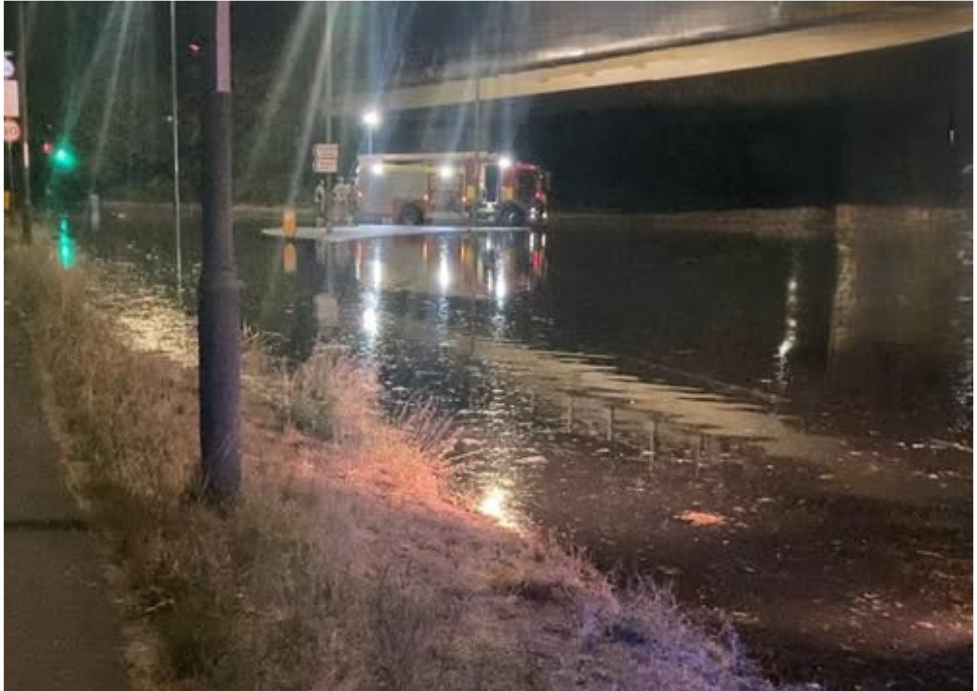A nighttime scene shows a flooded road beneath an overpass. A fire engine with its lights on is parked in the water, and a few emergency responders stand nearby.