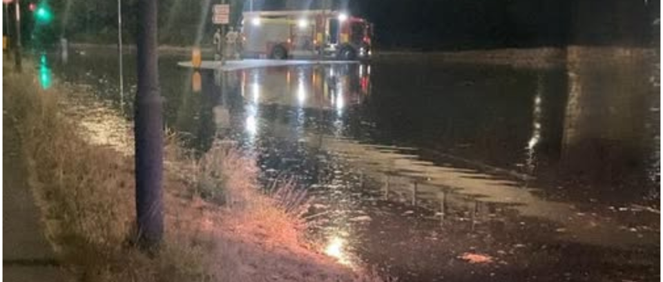 A nighttime scene shows a flooded road beneath an overpass. A fire engine with its lights on is parked in the water, and a few emergency responders stand nearby.