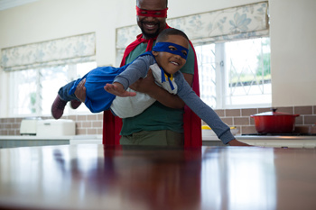Male foster carer holding a young boy wearing a superhero cape who is pretending to fly with his arms stretched out in front of him