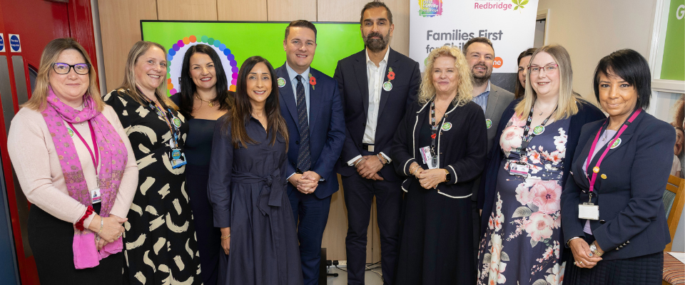 Group of eleven professionally dressed adults standing indoors in front of a "Families First for Children" banner, posing for a photo at a community or government event hosted by Redbridge.