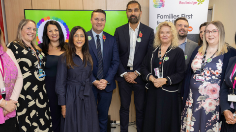 Group of eleven professionally dressed adults standing indoors in front of a "Families First for Children" banner, posing for a photo at a community or government event hosted by Redbridge.