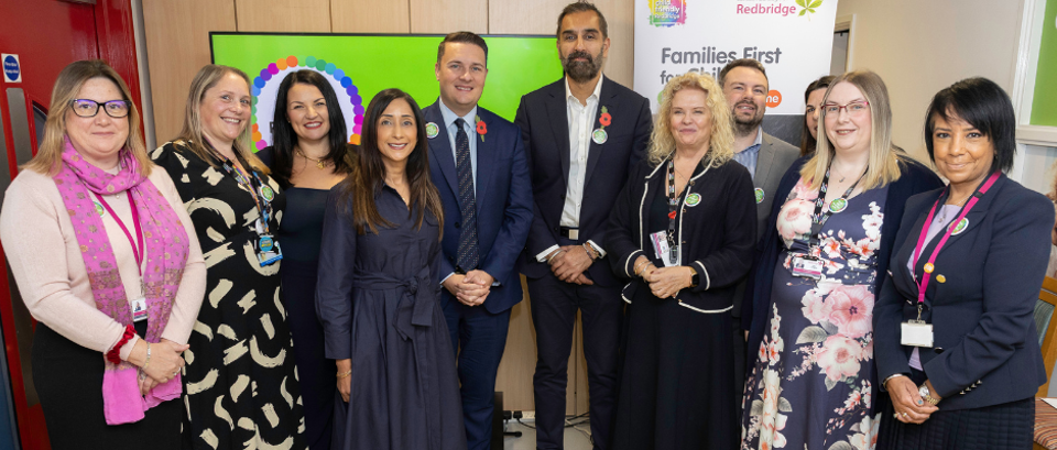 Group of eleven professionally dressed adults standing indoors in front of a "Families First for Children" banner, posing for a photo at a community or government event hosted by Redbridge.