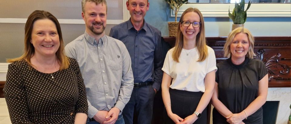 Five people standing indoors in a warmly lit room, smiling at the camera.